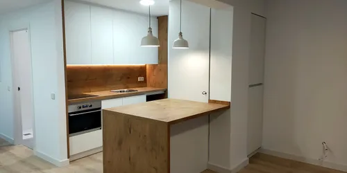 Cocina abierta al salón con muebles blancos, frente de roble con luz led y península de roble bajo dos lámparas blancas: cocina montada por Kitchenmania