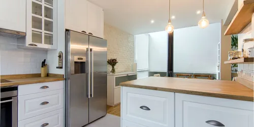 Cocina de puertas blancas con tirador cazoleta, encimera de madera de roble, frigorífico de acero y lámparas colgantes: cocina montada por Kitchenmania en Las Rozas