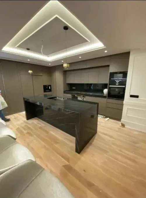 Cocina en tono gris topo con isla de piedra negra veteada, horno en columna y luz indirecta en el techo: cocina montada por Kitchenmania