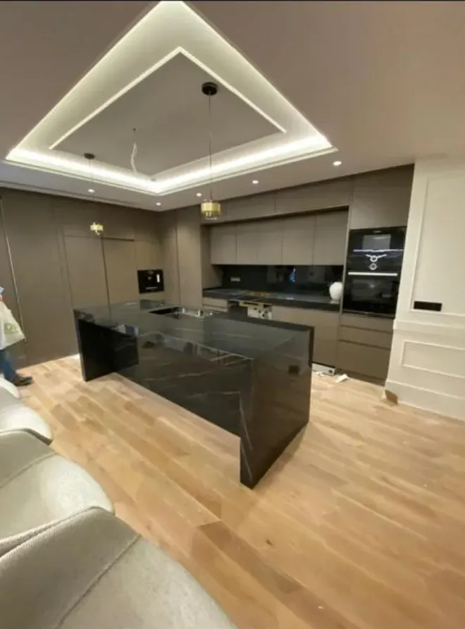 Cocina en tono gris topo con isla de piedra negra veteada, horno en columna y luz indirecta en el techo: cocina montada por Kitchenmania