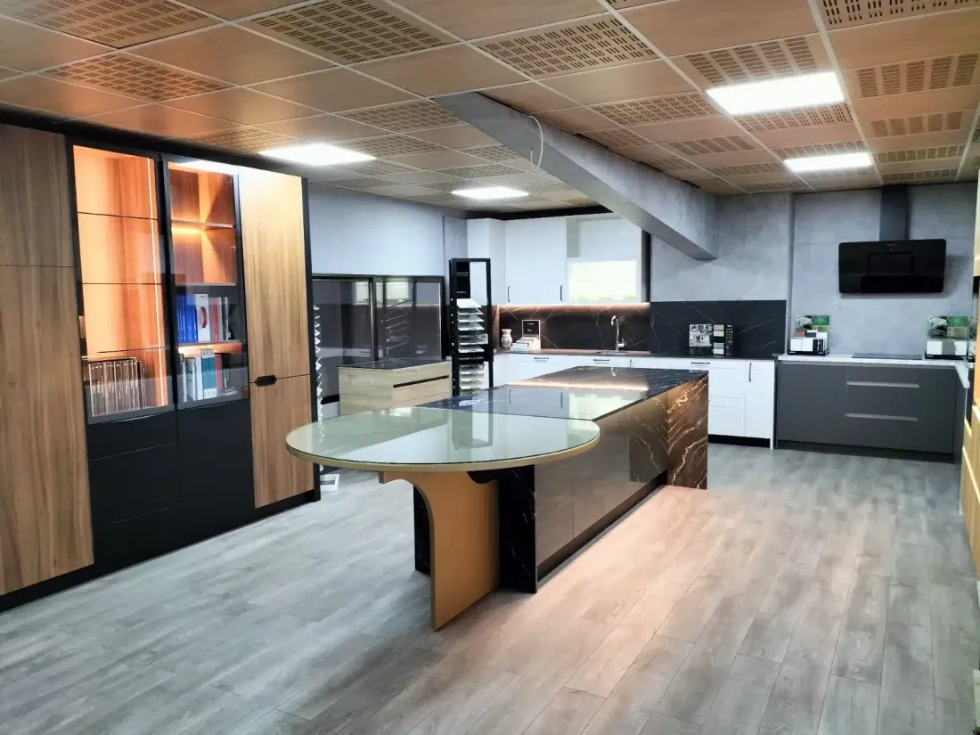 Isla con encimera de piedra oscura veteada, mesa redonda de cristal y columnas de madera con vitrinas iluminadas en la exposición de Kitchenmania en Illescas