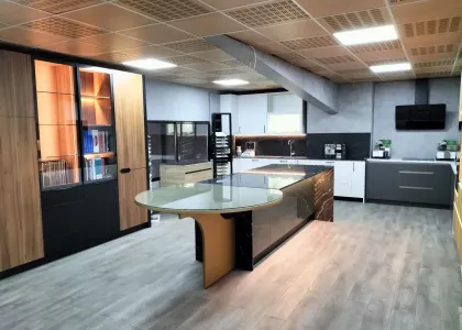 Isla con encimera de piedra oscura veteada, mesa redonda de cristal y columnas de madera con vitrinas iluminadas en la exposición de Kitchenmania en Illescas