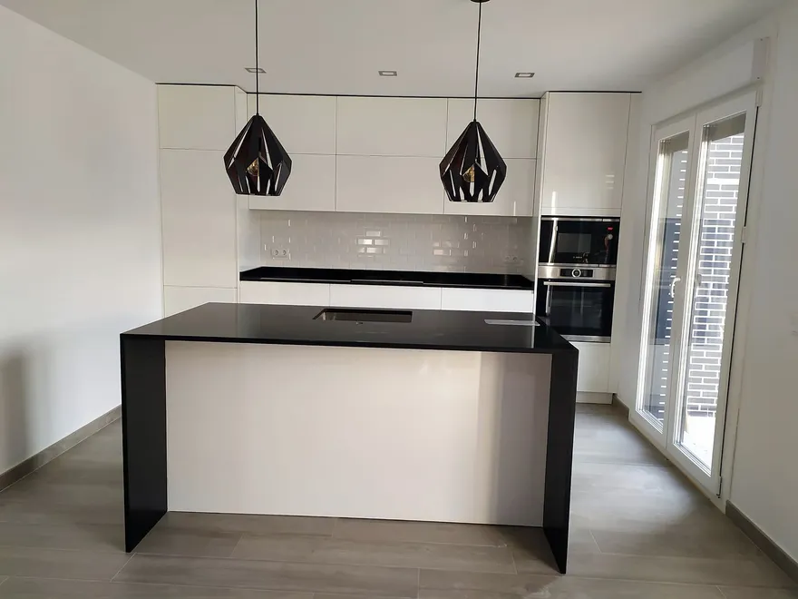 Isla de cocina con encimera negra y frente blanco bajo dos lámparas geométricas, muebles blancos y azulejo metro: cocina montada por Kitchenmania en Carranque (Toledo)