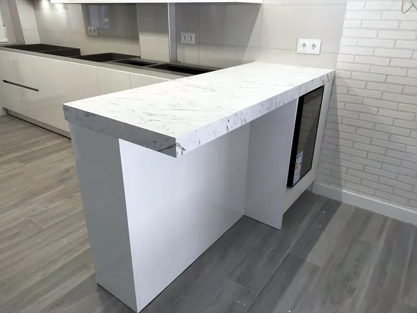 Península con encimera efecto mármol y vinoteca integrada junto a una pared de ladrillo blanco, muebles blancos en brillo: cocina montada por Kitchenmania en Alovera (Guadalajara)